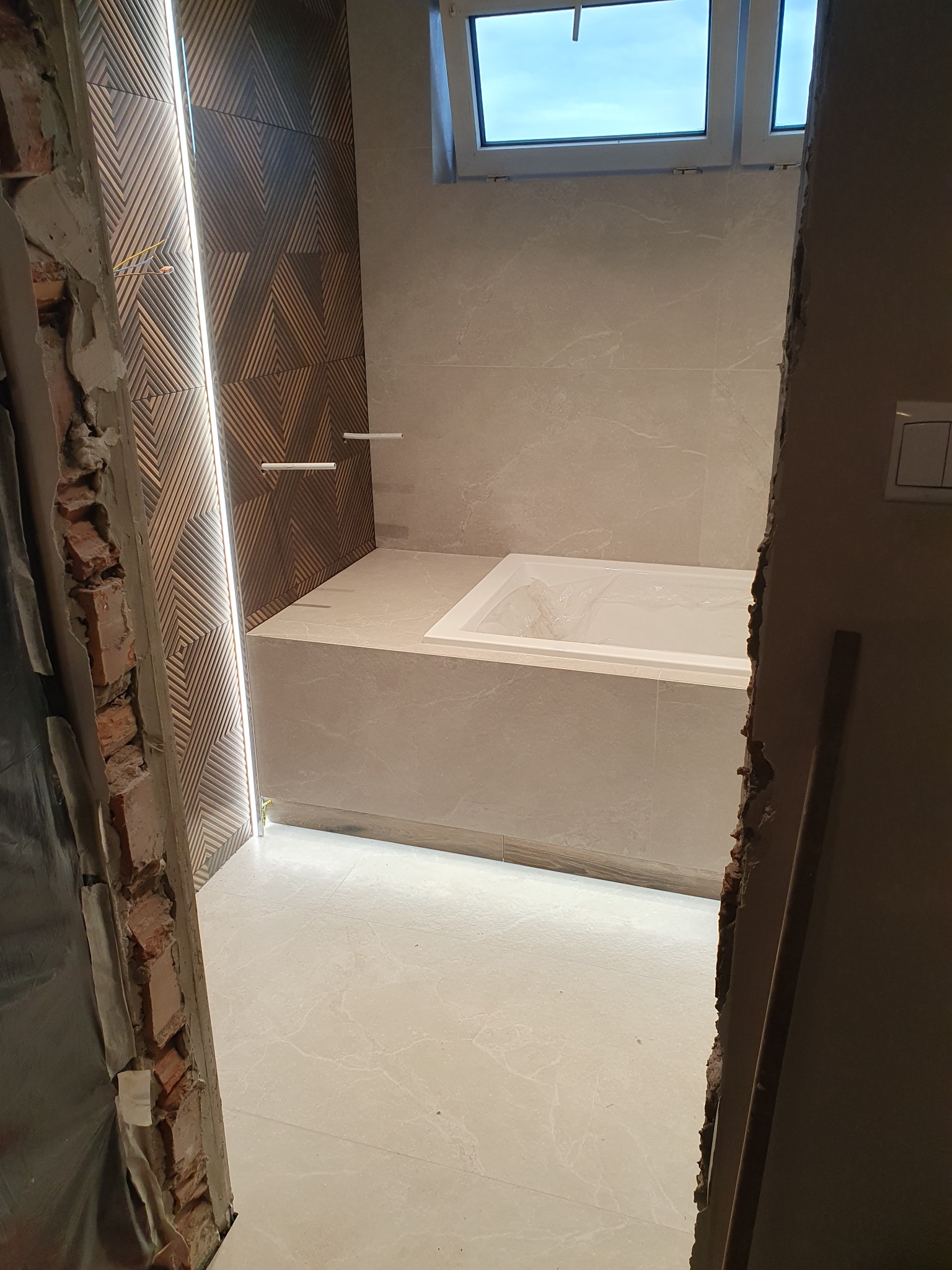 Remontowana łazienka z wanną w obudowie z płytek, dekoracyjną ścianą z podświetleniem LED i widocznymi fragmentami surowej ściany po demontażu ościeżnicy.