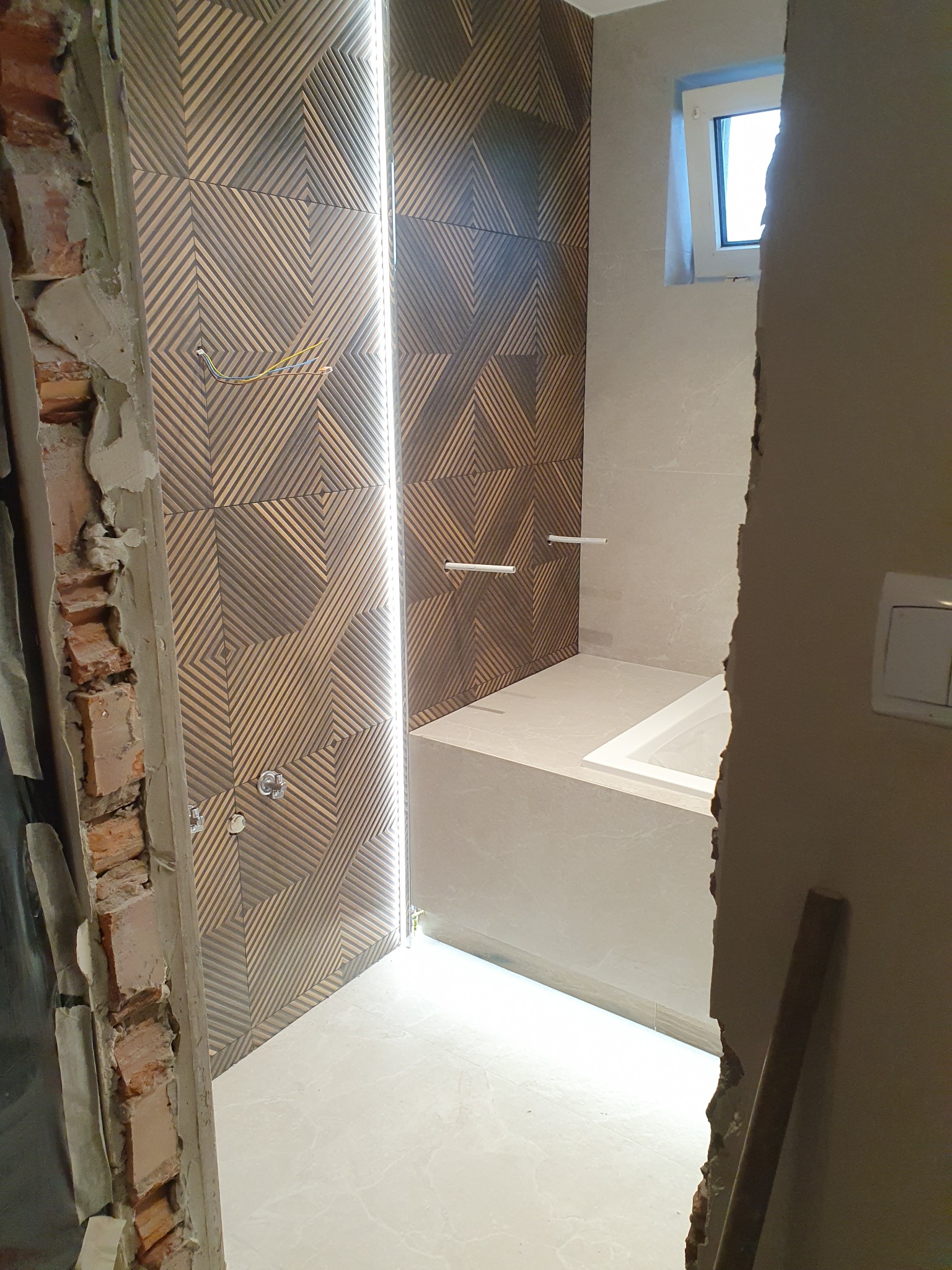 Łazienka w trakcie remontu, widoczne fragmenty ściany z cegły, ozdobne płytki na ścianie z podświetleniem LED, przygotowana instalacja wodna, biała wanna i jasna podłoga.