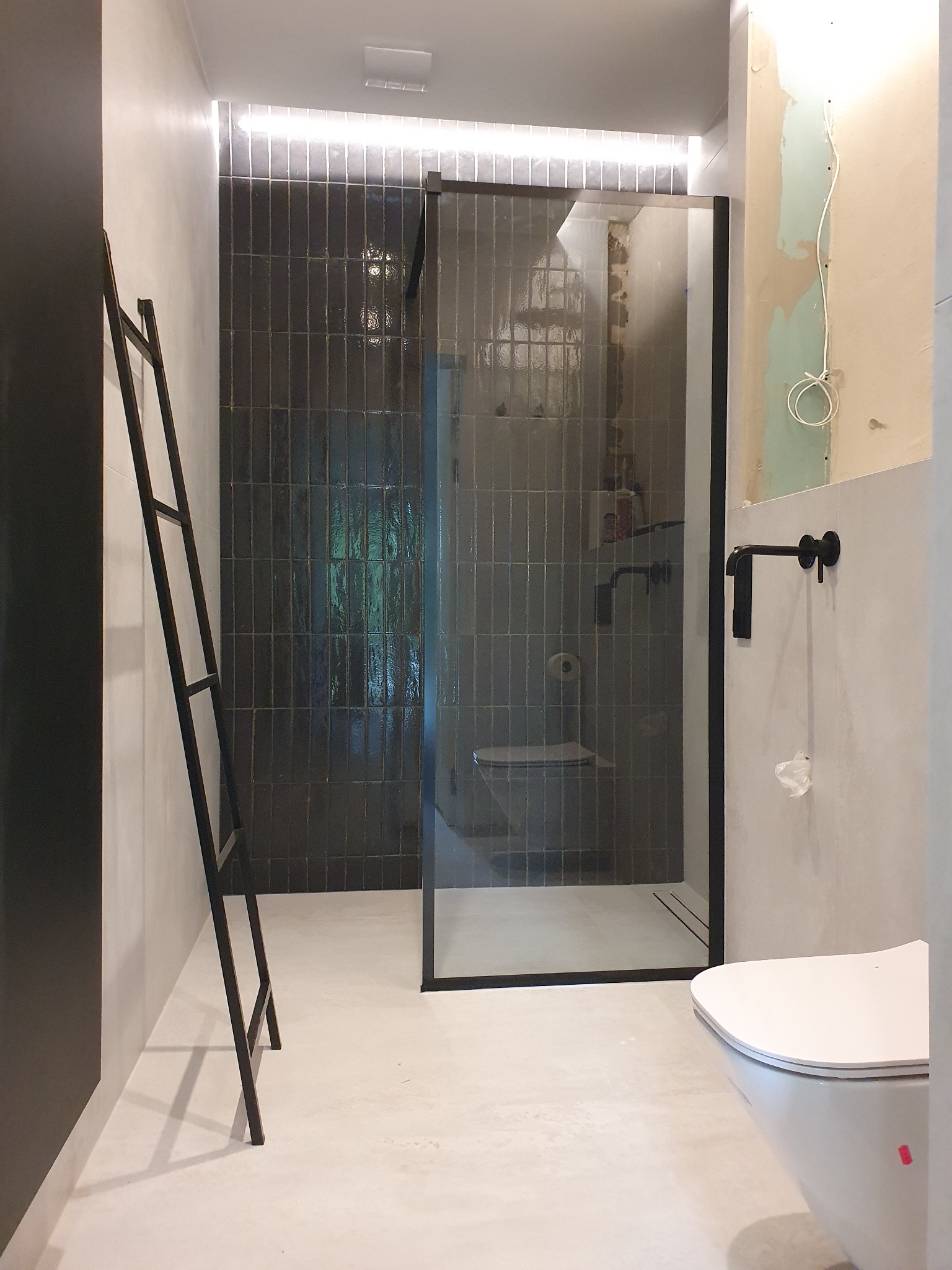 Łazienka w trakcie wykańczania: czarne, pionowe płytki w kabinie prysznicowej, czarna drabina na ręczniki, biała toaleta, surowe ściany w szpachli i widoczne przewody.