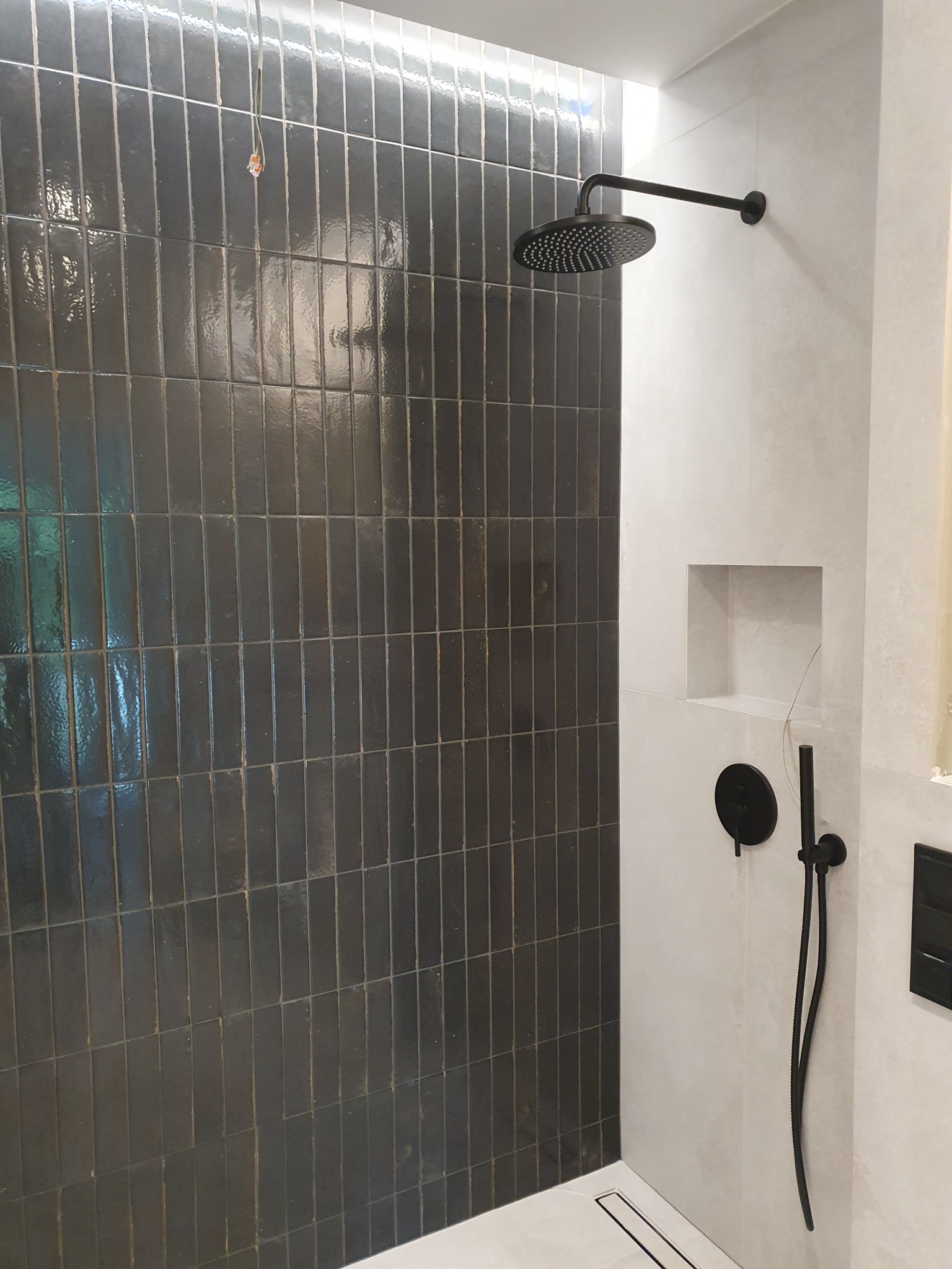 Nowoczesna kabina prysznicowa walk-in z czarną armaturą, ciemnymi, wąskimi płytkami ułożonymi pionowo i wnęką w ścianie.