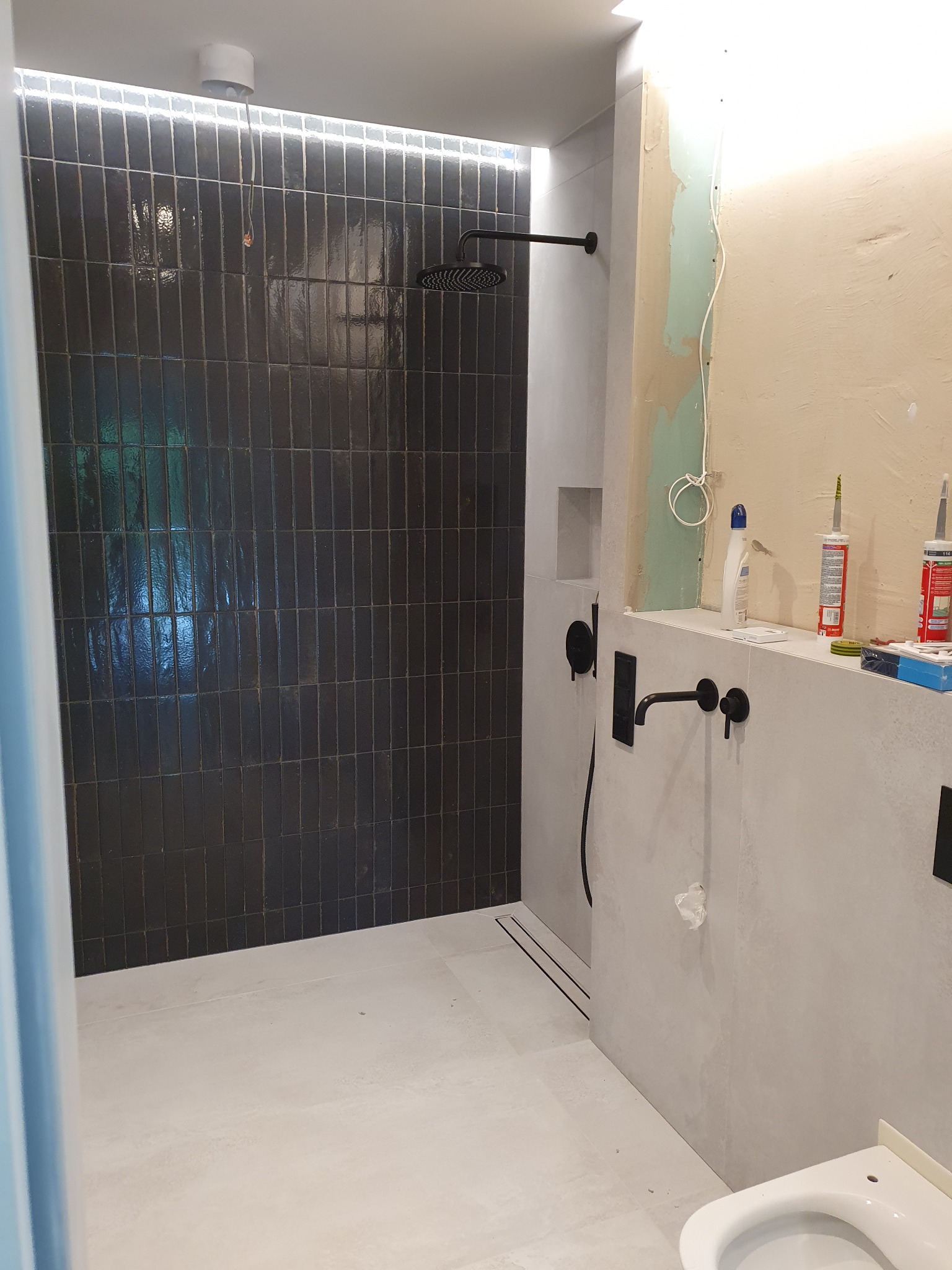 Wnętrze łazienki w trakcie wykańczania: czarne, pionowe płytki w kabinie prysznicowej z deszczownicą, surowy beton na ścianach, widoczne narzędzia i materiały budowlane, niedokończona instalacja...