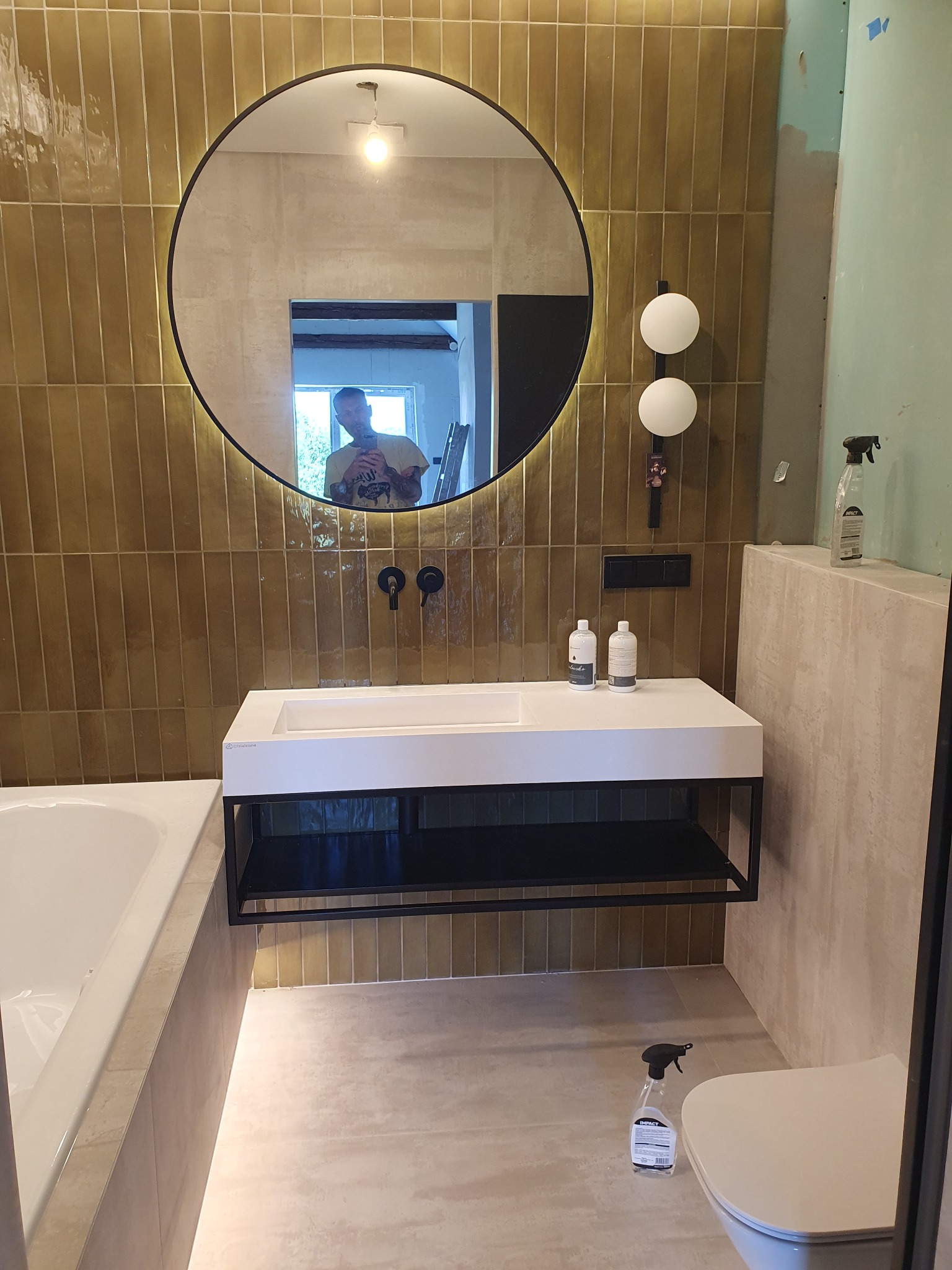 Nowoczesna łazienka w trakcie wykończenia: złote, wąskie płytki na ścianie, okrągłe lustro z podświetleniem, biała umywalka z czarnym stelażem, widoczna wanna i toaleta, fragment ściany w surowym...