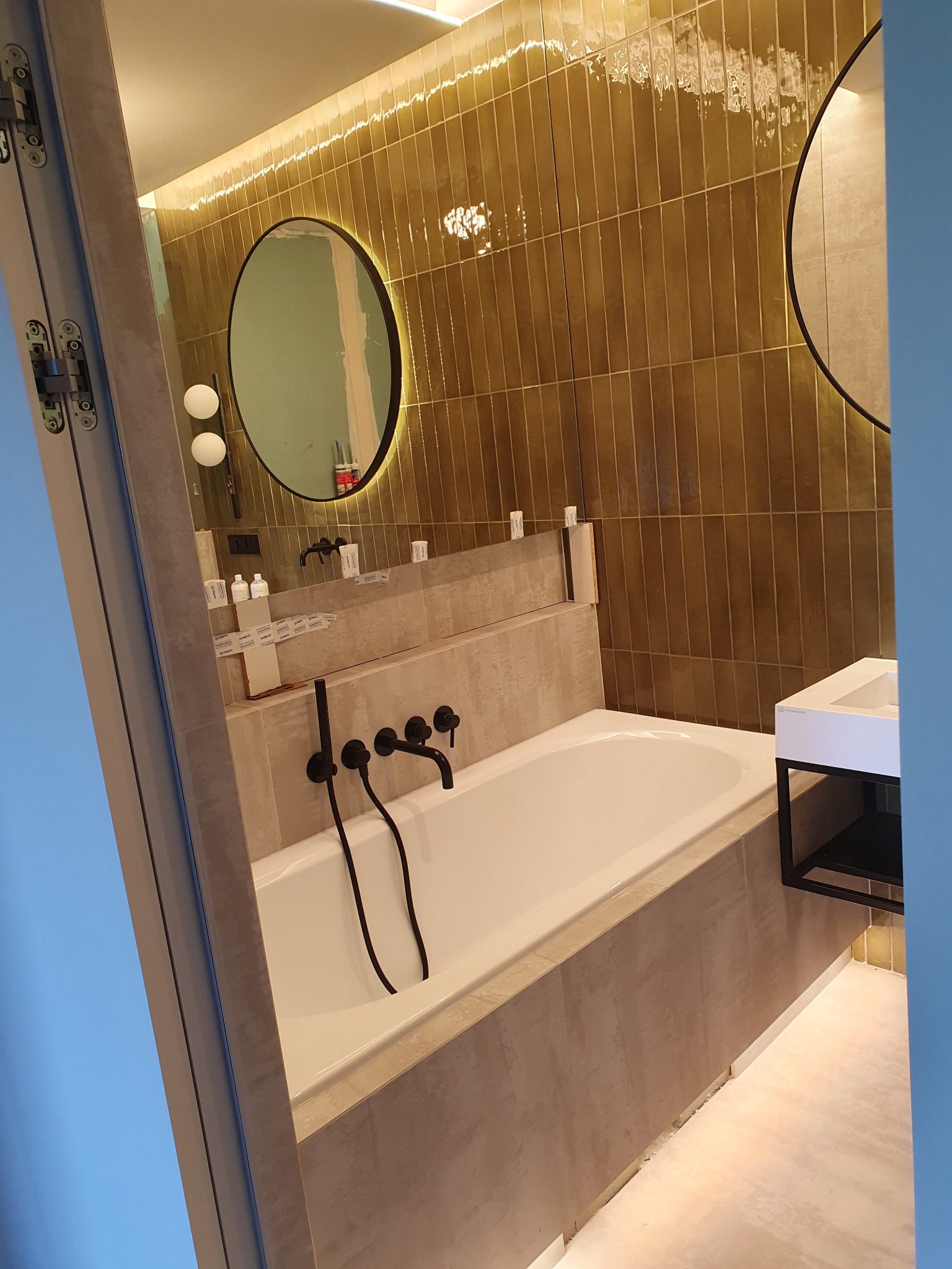 Nowoczesna łazienka z wanną obudowaną beżowymi płytkami, czarna armatura ścienna, okrągłe lustro z podświetleniem LED i pionowe płytki w odcieniach brązu na ścianie.