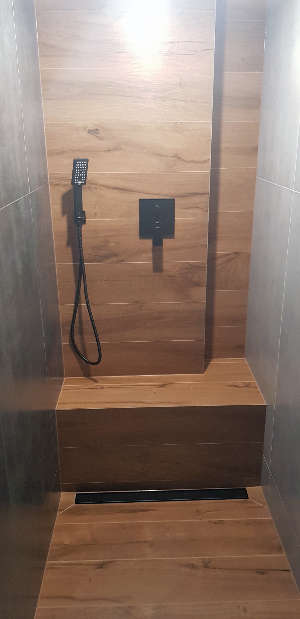 Nowoczesny prysznic walk-in z drewnopodobnymi płytkami, czarną armaturą i odpływem liniowym, minimalistyczny design.