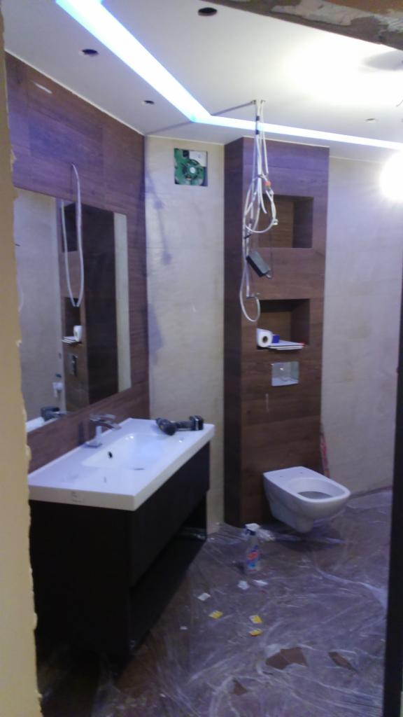 Łazienka w trakcie prac wykończeniowych: zamontowana umywalka z szafką, bidet, częściowo wykończone ściany z brązowymi panelami, widoczne narzędzia i folia ochronna na podłodze.