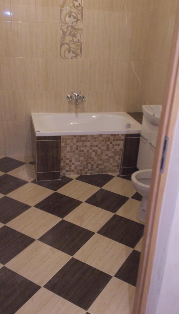 Wykończona łazienka z wanną obudowaną mozaiką, armaturą i szachownicą na podłodze w odcieniach beżu i brązu, widok zza framugi drzwi.