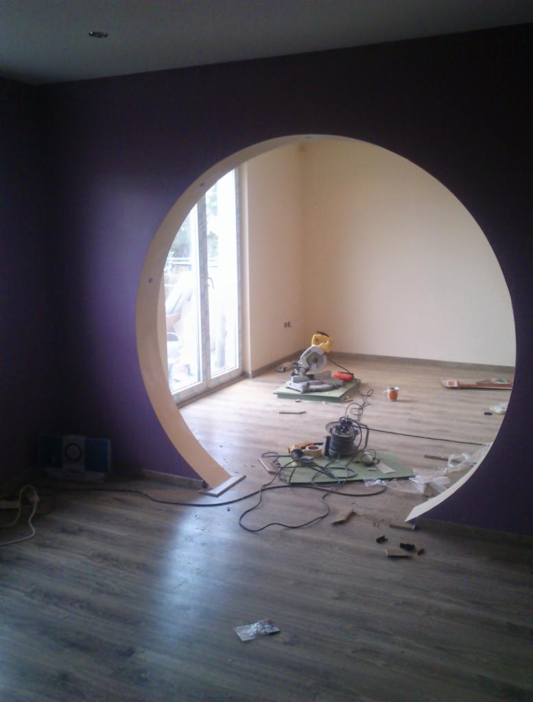 Wnętrze w trakcie wykończenia z okrągłym otworem w ścianie pomalowanej na fioletowo, widoczne narzędzia, kable i resztki materiałów na podłodze imitującej drewno.