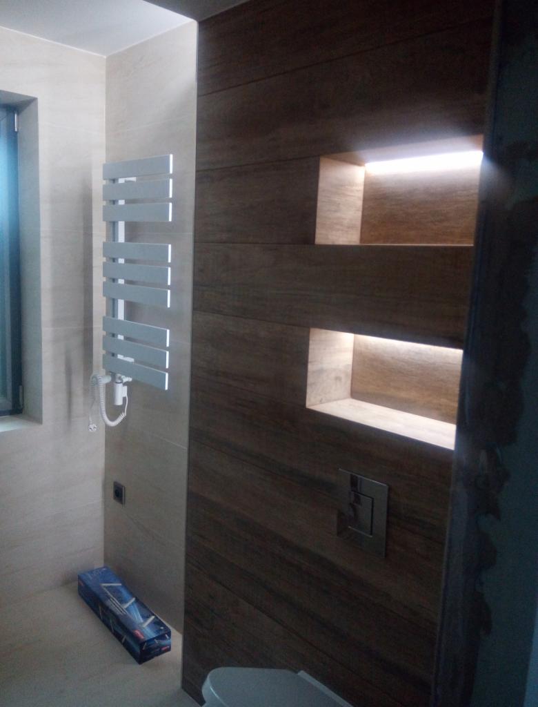 Nowoczesna łazienka z grzejnikiem drabinkowym, drewnopodobną okładziną ściany z podświetlanymi wnękami i częściowo widoczną toaletą. Na podłodze leży pudełko po produkcie.