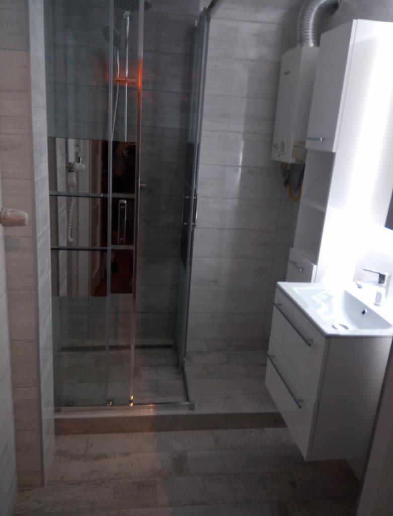 Nowoczesna łazienka z kabiną prysznicową ze szklanymi drzwiami przesuwnymi, białą szafką z umywalką i wiszącym piecykiem gazowym.