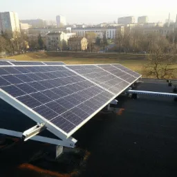 Panele słoneczne zamontowane na płaskim dachu budynku z widokiem na miejski krajobraz w tle.