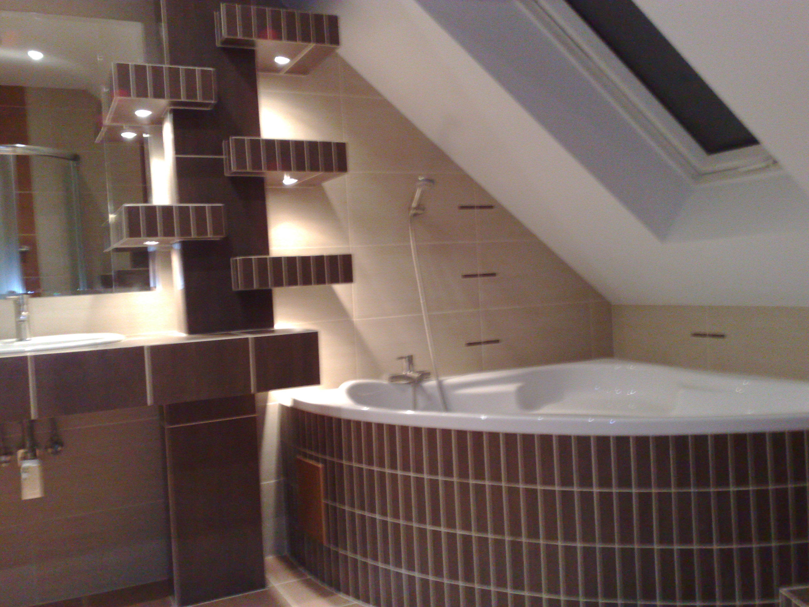 Wykończona łazienka na poddaszu w Jastrzębiu Zdroju, z narożną wanną obudowaną brązowymi płytkami, umywalką z szafką w tym samym kolorze oraz dekoracyjnymi półkami z oświetleniem.