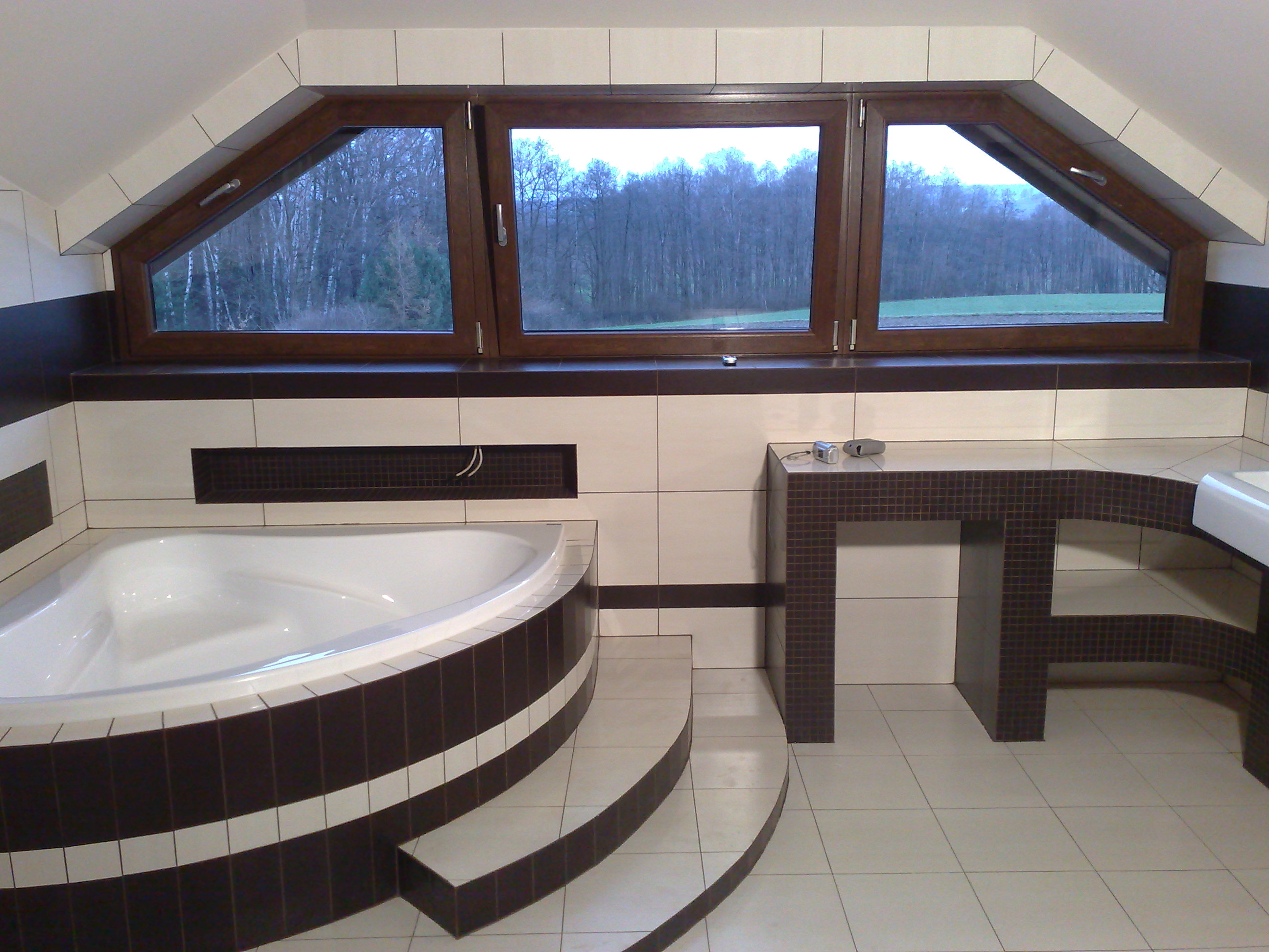 Wykończona łazienka na poddaszu z wanną narożną obudowaną brązowo-białymi płytkami, umywalką na blacie z mozaiki i oknem dachowym z widokiem na las.