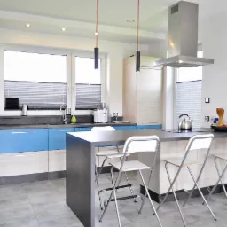 Nowoczesna kuchnia z jasnymi frontami szafek, błękitnymi akcentami, szarym blatem i wyspą z trzema białymi krzesłami barowymi, oświetlona wiszącymi lampami z czerwonymi kablami.