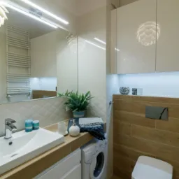 Jasna łazienka z białymi szafkami, drewnianym blatem pod umywalką, pralką i toaletą podwieszaną, oświetlona lampami sufitowymi i listwą LED