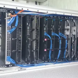 Rząd serwerów w szafie rack z niebieskimi kablami Ethernet, widoczne diody LED i etykiety na urządzeniach.