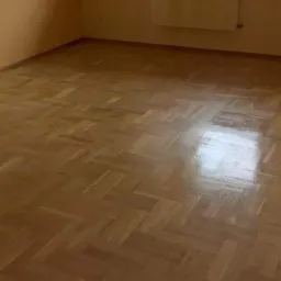 Parkiet ułożony w jodełkę, świeżo po lakierowaniu z widocznym odbiciem światła, na tle ściany w kolorze beżowym.