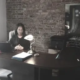 Kobieta siedzi za biurkiem w gabinecie z ceglaną ścianą, laptopem, lampką i dokumentami.