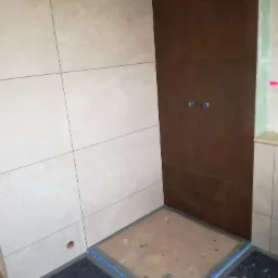 Niedokończona kabina prysznicowa z brązowymi i beżowymi płytkami, widoczne przygotowane podłoże pod brodzik i otwór na gniazdko elektryczne w ścianie