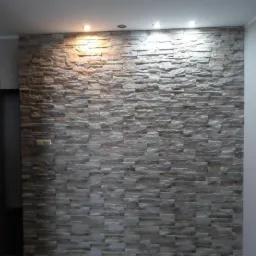 Ściana wykończona dekoracyjnym kamieniem w odcieniach szarości i beżu, podświetlona trzema punktowymi lampami sufitowymi, z widoczną podłogą z paneli imitujących drewno.