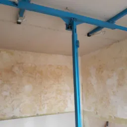 Niebieski podnośnik do płyt gipsowo-kartonowych, ustawiony w pomieszczeniu z częściowo przygotowanymi ścianami do dalszych prac wykończeniowych, widoczne ślady po starej farbie i nowe płyty gipsowe.