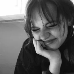 Portret młodej kobiety z przymkniętymi oczami, opierającej brodę na dłoni, w czarno-białej tonacji, z widocznymi piegami i kolczykiem w uchu, w tle okno.