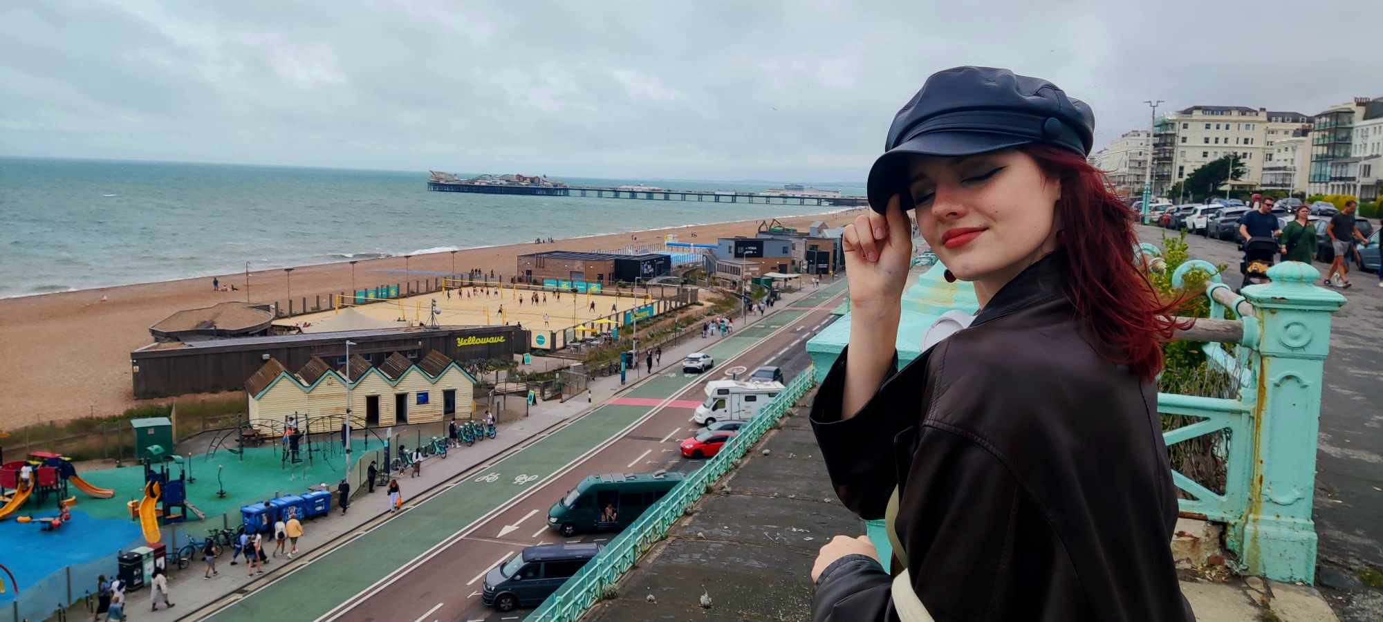 Kobieta w czapce z daszkiem pozuje na tle plaży z molo w Brighton, Anglia, widok z góry na ulicę z ruchem samochodowym i pieszym.