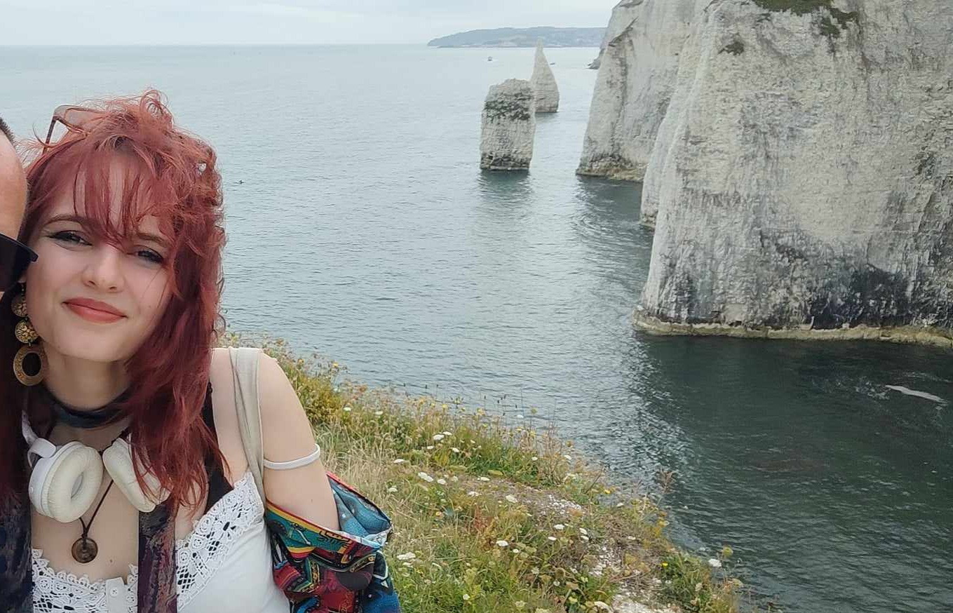 Portret osoby z rudymi włosami na tle formacji skalnych w morzu, widoczne klify i woda.