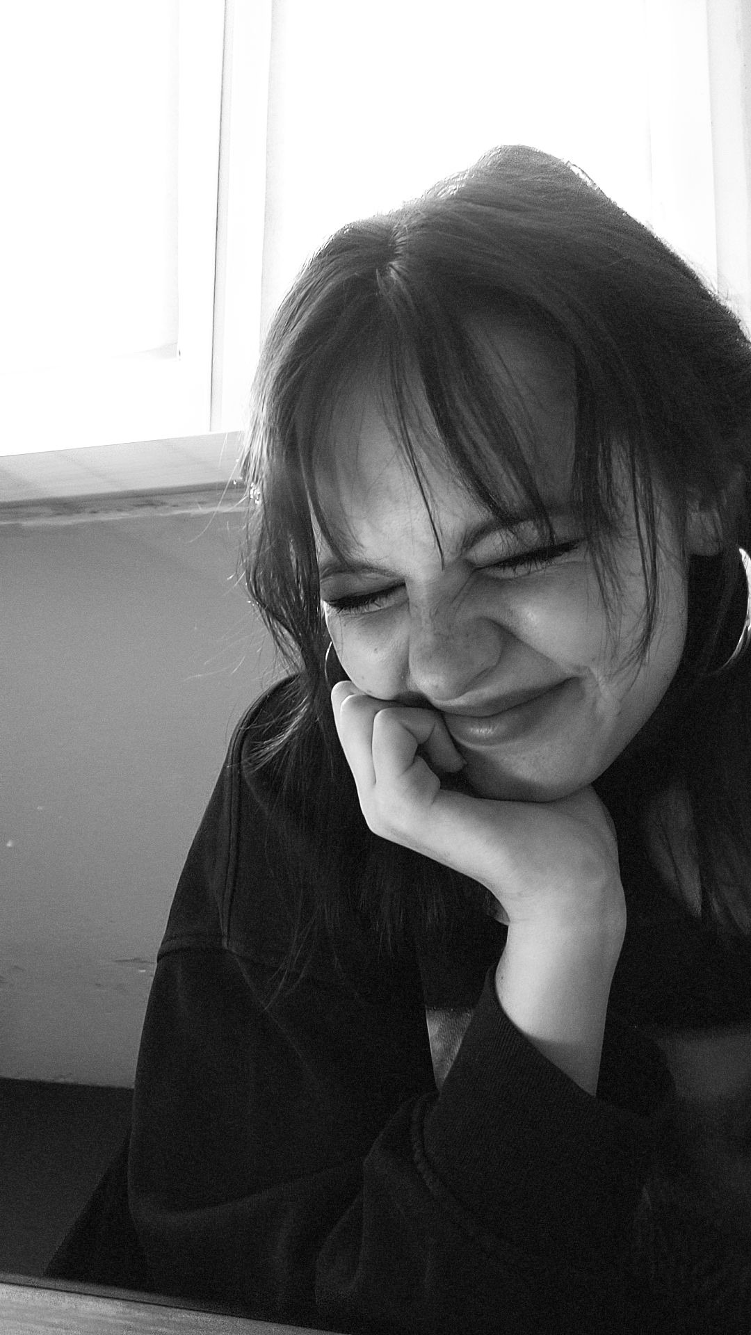 Portret młodej kobiety z przymkniętymi oczami, opierającej brodę na dłoni, w czarno-białej tonacji, z widocznymi piegami i kolczykiem w uchu, w tle okno.