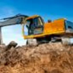 Żółta koparka na hałdzie ziemi, widok z dołu na tle błękitnego nieba.