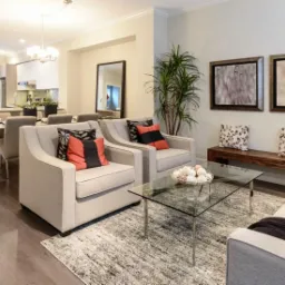 Jasne, przestronne wnętrze salonu z drewnianą podłogą, szarymi fotelami z kolorowymi poduszkami, szklanym stolikiem kawowym i widokiem na jadalnię oraz kuchnię w tle.