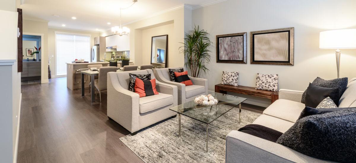 Jasne, przestronne wnętrze salonu z drewnianą podłogą, szarymi fotelami z kolorowymi poduszkami, szklanym stolikiem kawowym i widokiem na jadalnię oraz kuchnię w tle.