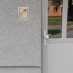 Szary domofon z klawiaturą numeryczną zamontowany na elewacji budynku obok szarych drzwi wejściowych z szybą, widok z zewnątrz.