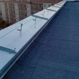 Metalowa listwa odgromowa z uchwytami zamontowana na płaskim dachu budynku wielorodzinnego o zmierzchu.