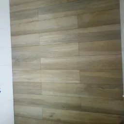 Ściana wyłożona podłużnymi płytkami ceramicznymi imitującymi drewno w odcieniach beżu i brązu, ułożonymi poziomo, z fragmentem białej ściany z lewej strony.