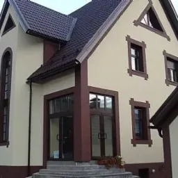 Dwukondygnacyjny dom z beżową elewacją wykończoną brązowymi detalami, ciemnobrązowy dach, widok z zewnątrz na wejście i okna.