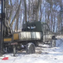 Wiertnica zamontowana na samochodzie terenowym w zimowym krajobrazie, przygotowana do wiercenia, z widocznym świdrem i czerwonym pojemnikiem na śniegu.