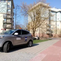 Srebrny samochód z naklejkami firmy na tle bloków mieszkalnych i chodnika z kostki brukowej.