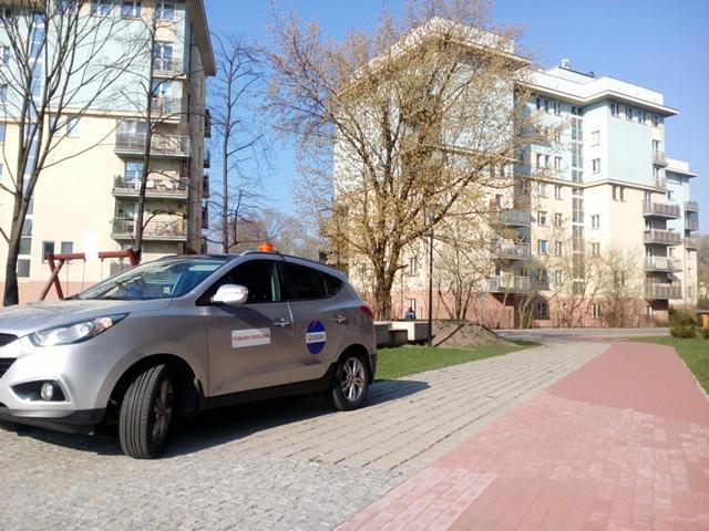 Srebrny samochód z naklejkami firmy na tle bloków mieszkalnych i chodnika z kostki brukowej.