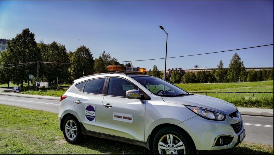 Srebrny samochód z napisem 'Pomiary Drogowe' i pomarańczowymi światłami ostrzegawczymi na dachu, zaparkowany przy drodze, w tle zielony krajobraz z zaporą wodną.