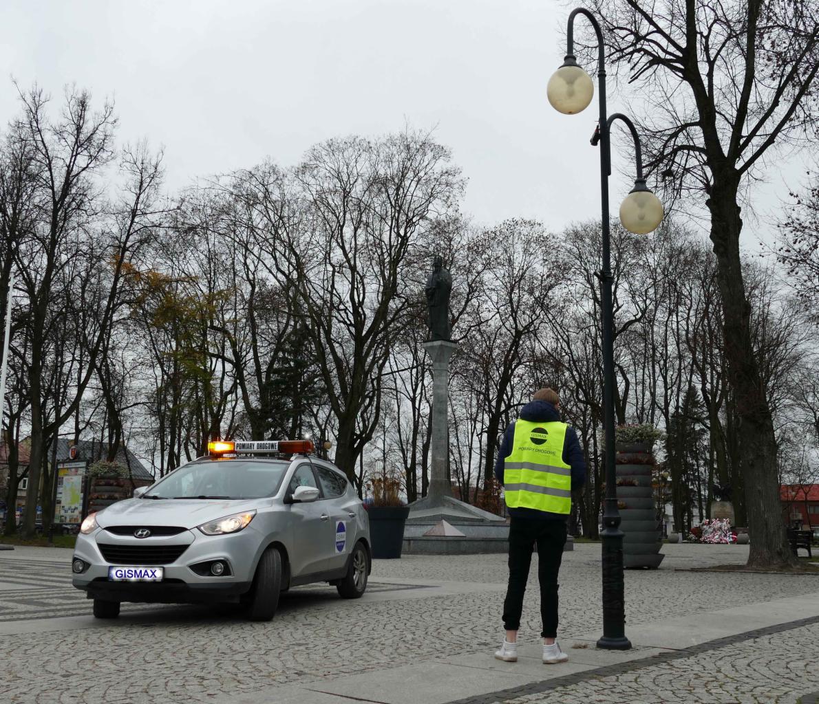Srebrny samochód z napisem 'Pomiary Drogowe' na dachu, stojący na placu z kostki brukowej, obok mężczyzna w żółtej kamizelce odblaskowej, w tle drzewa i pomnik.