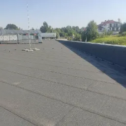 Szary, płaski dach budynku komercyjnego z widocznymi elementami wentylacyjnymi, anteną i obróbkami blacharskimi, w tle budynki mieszkalne i znak McDonald's.