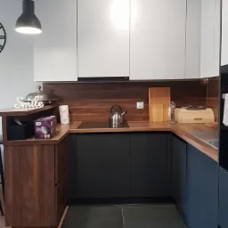 Nowoczesna kuchnia z białymi szafkami górnymi, ciemnymi szafkami dolnymi i drewnianym blatem, czajnik na płycie indukcyjnej, widoczny narożnik z barkiem i dekoracyjny napis 'Smile'.