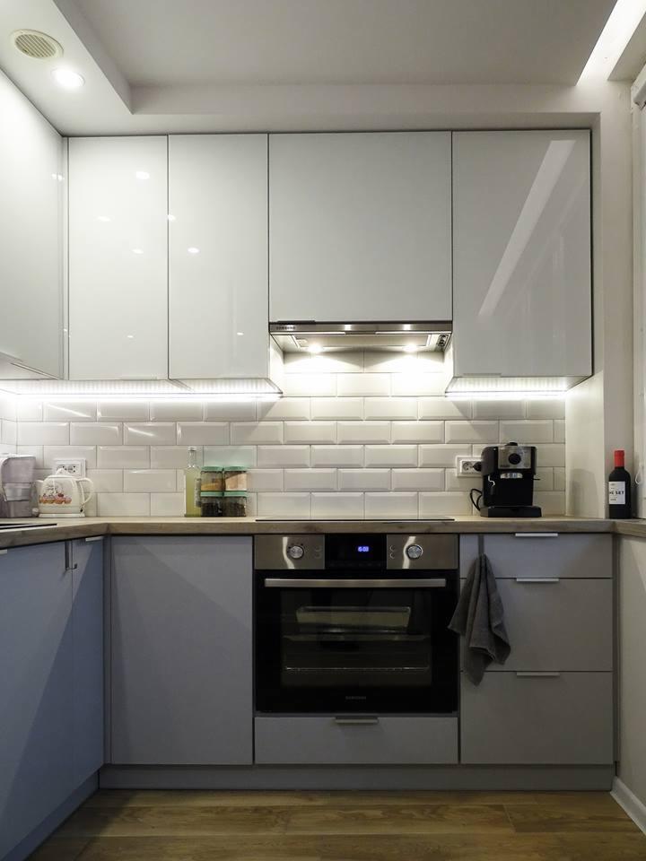 Nowoczesna kuchnia z białymi, lakierowanymi szafkami górnymi, szarymi szafkami dolnymi i oświetleniem LED pod szafkami, widoczny ekspres do kawy i butelka wina.