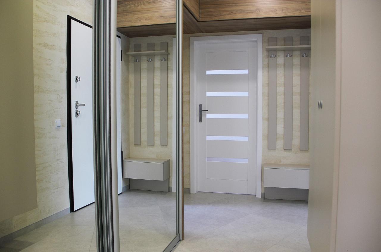 Nowoczesny przedpokój z przesuwną szafą lustrzaną, białą stolarką drzwiową z poziomymi przeszkleniami oraz panelami ściennymi z wieszakami i siedziskiem.