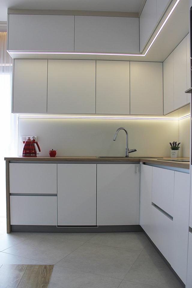 Nowoczesna, jasna kuchnia z białymi szafkami, drewnianym blatem, oświetleniem LED pod szafkami wiszącymi, czerwonym czajnikiem i jabłkiem na blacie oraz szarą podłogą z płytek.