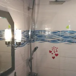 Wykończona łazienka z biało-niebieskimi płytkami z motywem fal, deszczownicą i naklejkami w kształcie serc na ścianie. Widoczny fragment lustra z oświetleniem oraz akcesoria łazienkowe przy wannie.