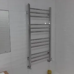 Chromowany drabinkowy grzejnik łazienkowy zamontowany na białej, błyszczącej ścianie z płytek ceramicznych. Widoczne przyłącza grzejnika.