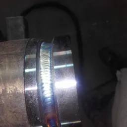 Spoina na rurze ze stali nierdzewnej z widocznymi efektami termicznymi, wykonana metodą TIG, z fragmentem ubrania roboczego w tle.