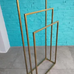 Złoty, metalowy stojak na ręczniki o geometrycznym kształcie, na tle turkusowej ściany imitującej kamień i szarej podłogi z płytek.