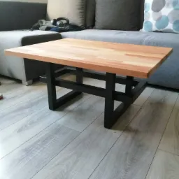 Stolik kawowy z drewnianym blatem i czarną metalową podstawą o geometrycznym kształcie, ustawiony na panelach podłogowych w odcieniach szarości, z widoczną częścią szarej sofy i poduszek w tle.